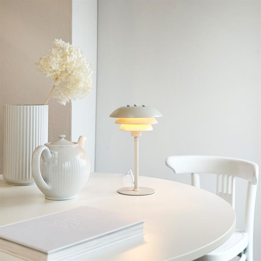 Weiße Tischlampe spendet sanftes, warmes Licht auf einem runden weißen Tisch; daneben steht eine Teekanne aus Porzellan, eine hohe, geriffelte Vase mit einer Trockenblume und ein weißer Stuhl.