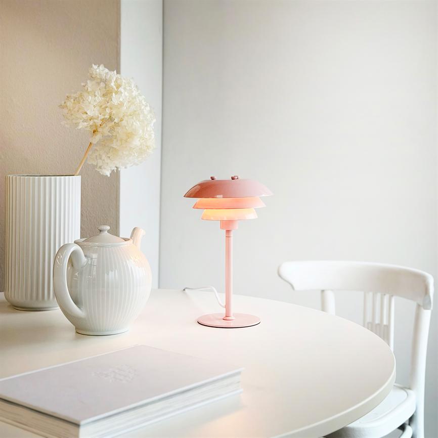 Eine rosa Tischlampe leuchtet warm auf einem runden weißen Tisch, umgeben von einer weißen Teekanne, einer hohen Vase mit Trockenblumen und einem leeren weißen Stuhl in einem neutralen Raum.